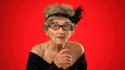 Portrait of funny looking angry upset frustrated elderly senior old woman saying shh, quiet, with wrinkled skin and grey hair on red background.