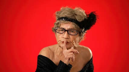 Portrait of funny looking angry upset frustrated elderly senior old woman saying shh, quiet, with wrinkled skin and grey hair on red background.