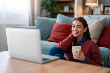 A cheerful caucasian woman participates in a virtual meeting from her cozy living room, comfortably dressed with a mug in hand, using her laptop.