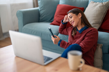 A smiling Caucasian woman efficiently manages her tasks, browsing on her laptop while texting on her smartphone, comfortably seated on a living room sofa with a mug nearby.