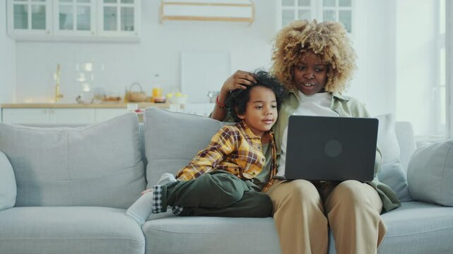 Young Black mother hugging little son and discussing something on laptop screen with him, both sitting on couch in modern minimalist apartment