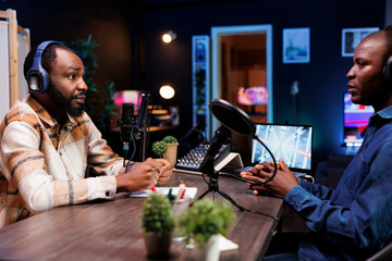 Black man hosting radio program is having live conversation with male guest, recording the encounter for audience to listen. Influencer making a podcast with journalist using audio equipment.