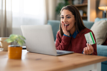 A cheerful Caucasian woman engages in online shopping, holding a credit card while using her laptop in a cozy, modern living room.