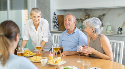 Happy carefree older man and women gathering around kitchen table, sipping alcoholic drinks with...