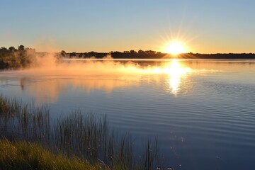 Obraz premium Tranquil Sunrise Over Misty Lake with Grassy Shoreline and Golden Reflection