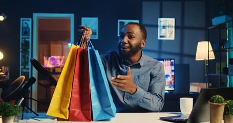 Content creator holding shopping bags, filming haul video in personal apartment studio. Internet star unpacking clothing items he bought, reviewing fashion stores merchandise