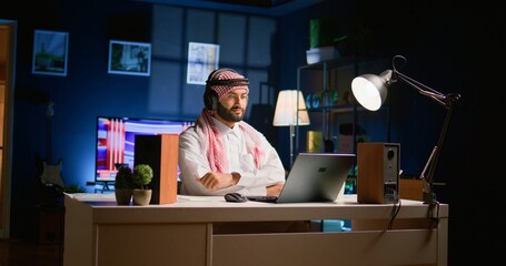 Arab man with wireless headphones at home in living room, watching educational videos on laptop....