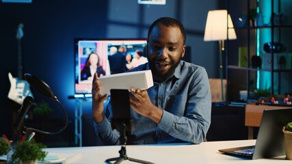 Tech content creator filming packaged product unboxing in neon lights personal home studio using professional equipment. Influencer showing sponsoring partner merchandise, urging subscribers to buy it