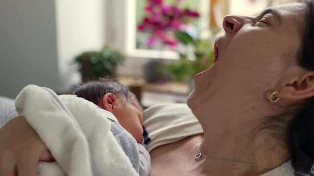 Mother yawning while holding her newborn baby close, illustrating the exhaustion and tender moments of early parenthood, emphasizing the bond and love shared despite the challenges