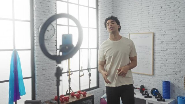 Young hispanic man in a gym recording a video with a ring light and smartphone, showing fitness exercises with gym equipment in the background.
