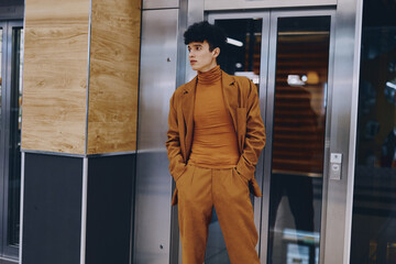 Stylish young man in a brown suit and turtleneck standing confidently near an elevator in a modern urban setting