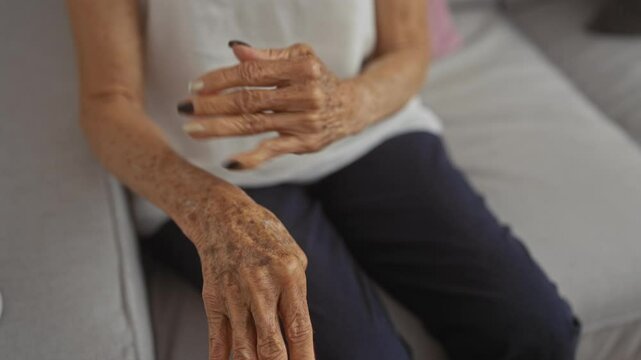 An elderly woman sits in her living room, focusing on her hands which show visible signs of aging and have well-manicured nails.
