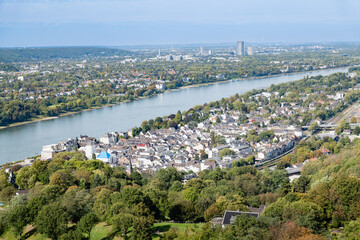 Fototapeta premium Panoramic view of the town and the meandering Rhine River, taken from Schloss Drachenburg. The landscape is dotted with houses, greenery, and distant city buildings under a clear blue sky.
