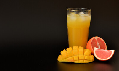 Grapefruit-mango juice with ice in a tall faceted glass on a black background, next to chopped ripe fruits.