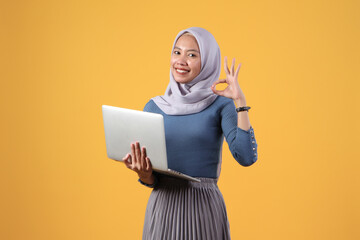 happy asian indonesian muslim woman holding laptop computer giving ok finger sign on isolated yellow background