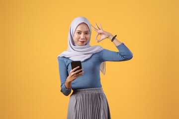 happy asian indonesian muslim woman holding smart phone giving ok finger gesture on isolated yellow...