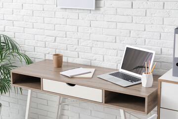 Workplace with laptop, stationery and notebooks near white brick wall in office