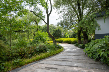 A serene and inviting outdoor space, well-maintained with manicured and trimmed hedges in urban parkland. A winding pathway leads through the park, with a variety of trees and shrubs