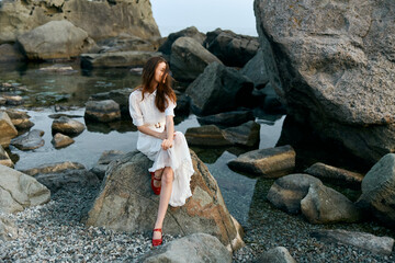 Serene woman in white dress with red shoes sitting on rocky shoreline by the ocean