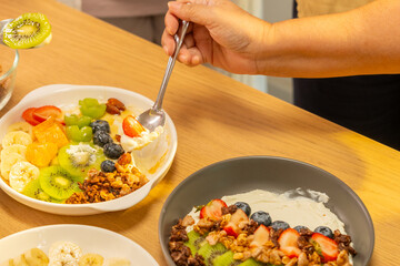 Group of Happy Asian senior women having breakfast together at home. Elderly retired woman friends enjoy healthy lifestyle making homemade greek yogurt with mixed fruit, nuts and honey in the kitchen.