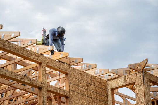 New residential home construction in framing stage, workman with hardhat and toolbelt on the roof rafters placing board with air vent holes between rafter frames
