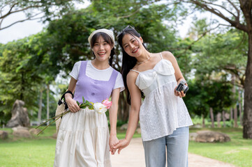 Two lovely Asian women are holding hands, strolling through a park, and smiling at the camera.