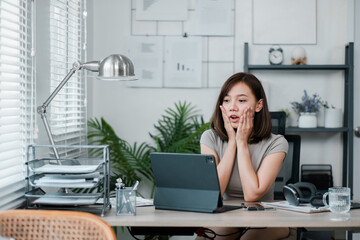 Young woman working from home office, looking surprised while using a tablet. Modern workspace with plants, shelves, and office supplies.