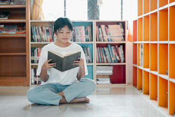 A young woman sits cross-legged on the floor, reading a book in a bright, modern library with colorful shelves and natural light.