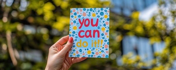 A person holding up an inspiring card that says "you can do it!" with colorful text, representing positive self-affirmation and positivity.