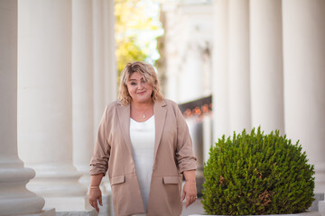 A beautiful fat middle aged woman walks around the city in a stylish image. The plus size model is dressed in a biege jacket and a white dress