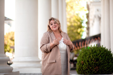 Fototapeta premium A beautiful fat middle aged woman walks around the city in a stylish image. The plus size model is dressed in a biege jacket and a white dress