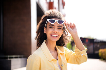 Photo of young joyful girl take off sunglasses with wavy hair student in yellow stylish jacket spending time outdoors sunny weather
