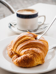 Breakfast with buttery croissant and a fork on a white plate, accompanied by a cup of white coffee in an outdoors cafe.