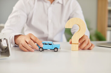 Male worker sitting at an office desk and holding a little blue toy automobile model and a wooden...