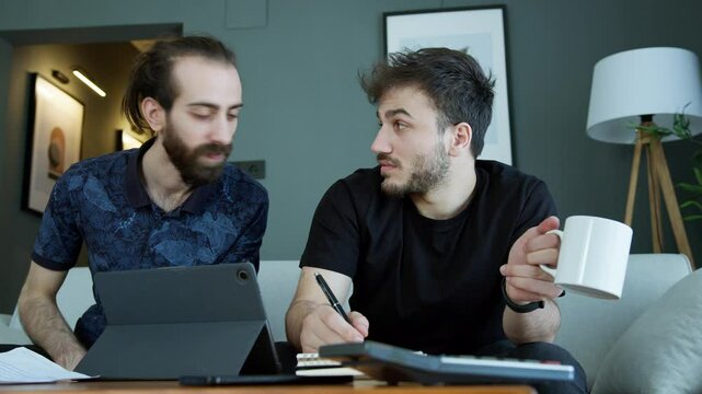 Two men are sitting on a couch, collaborating on a project. The bearded man with a displeased expression, critiques the work done his friend.