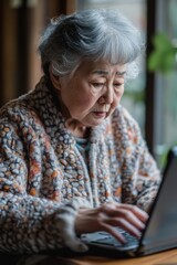 Japanese senior woman learning to use a laptop, fingers typing on the keyboard, hand on the mouse, focused on the screen.