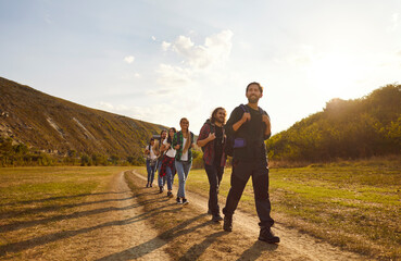 Young happy people tourists with large backpacks hiking in nature walking in a row in the mountains. Smiling friends having trekking tour outdoors. Adventure, travel and tourism concept.