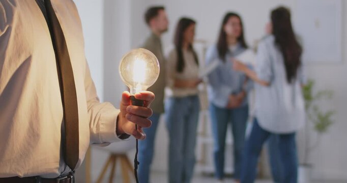 Man holding light bulb bright lamp as new idea, thinking alone, problem solution, worried puzzled manager pondering question or busy solving business issues at workplace, team at background, close up
