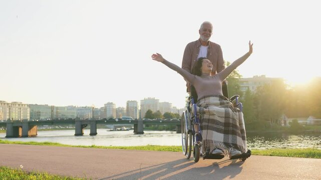 Senior woman in wheelchair walking with caregiver old man on road. Family couple man husband supporting taking care of paralyzed woman wife in chair for people with disability outdoor. Rehabilitation
