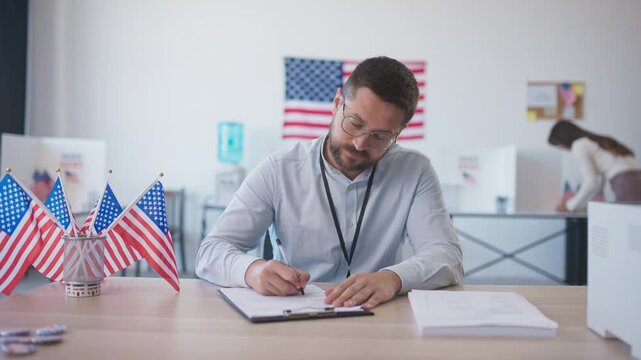 Caucasian man calmly sitting at desk and filling in information on piece of paper. Miniature flags of US located on table. Woman in blurred background voting for president or congress member.