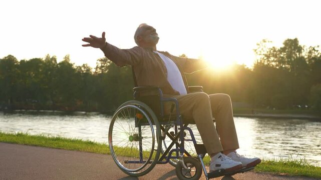 Happy senior man in wheelchair raised hands enjoying freedom walking on road in city park outdoor. Old happy man in chair for people with disability has a hope. Disability disease rehabilitation
