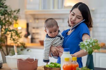 A woman is talking on her cell phone while holding a baby. The kitchen is filled with various items such as bowls, bottles, and vases