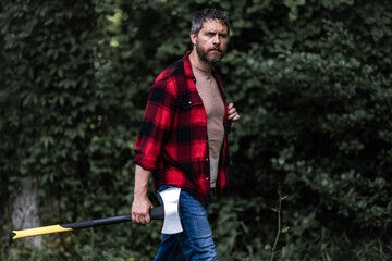 Fototapeta premium Lumberjack in shirt with heavy axe. Strong man work in forest. Lumber jack men in forest with axe. Lumberjack carries axe. Deforestation. Handsome man with axe. Lumberjack in the woods with an ax.