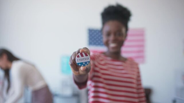 African American woman in blurred vision shoving to camera round pin saying "I voted". People voting in background. American flag hanging on wall. Presidential Elections in United States of America.