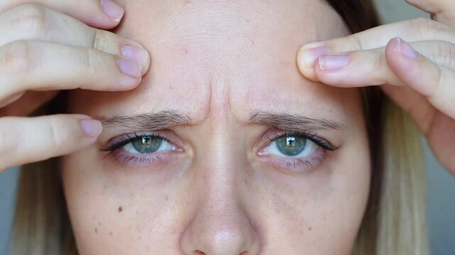 Close up of a young caucasian woman frowning touching the forehead showing wrinkles on her face on a grey background. The inter-brow wrinkle on the forehead. Skin care. Beauty concept