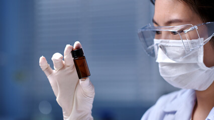 The female scientist in protective eyeglasses holding an amber experiment sample storage vial in...