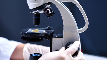 Laboratory concept template photo to design advertising pic, close-up view of a scientist's hand wearing white gloves adjusting a microscope to observe yellow cream placed under the microscope