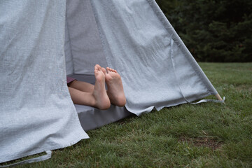 Bare feet of little girl sitting in textile toy wigwam in garden at summer day © Natalia Navodnaia