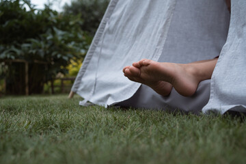 Bare feet of little girl sitting in textile toy wigwam in garden at summer day © Natalia Navodnaia