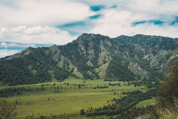 Lush green valleys contrast with the rocky peaks of the Altai Mountains on a cloudy day, showcasing the region's natural beauty and tranquility